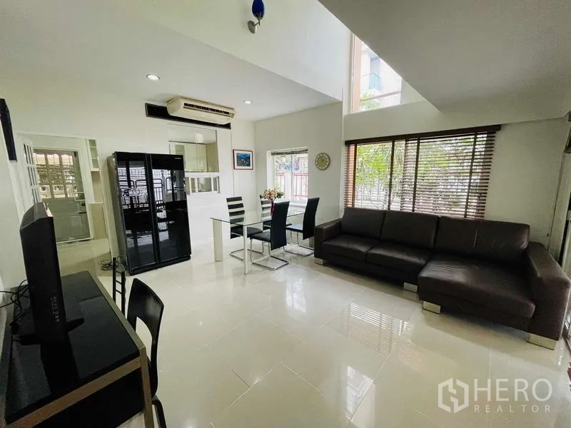 Detached House for rent in Watthana, Bangkok - Dining and family area with sectional sofa, glass dining set, and tall windows.