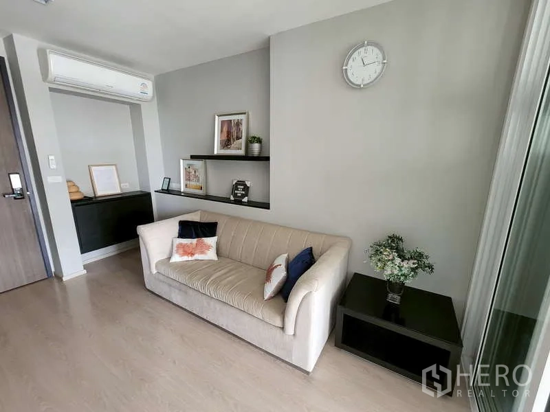 Condominium for sale - Cozy lounge area featuring a beige sofa, floating shelves with decor, wall clock and side table under an air conditioner.
