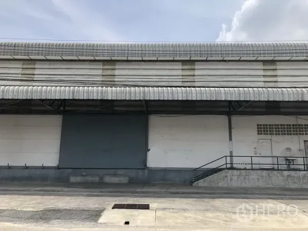 Warehouse for rent in Khlong Luang, Pathum Thani - Warehouse exterior with canopy roof, ramp access, and a large metal sliding door.