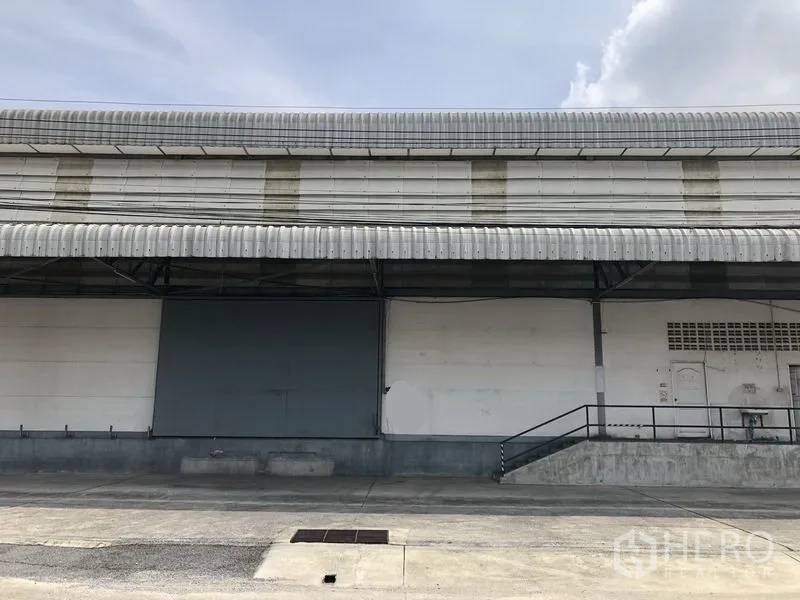Warehouse for rent in Khlong Luang, Pathum Thani - Warehouse exterior with canopy roof, ramp access, and a large metal sliding door.