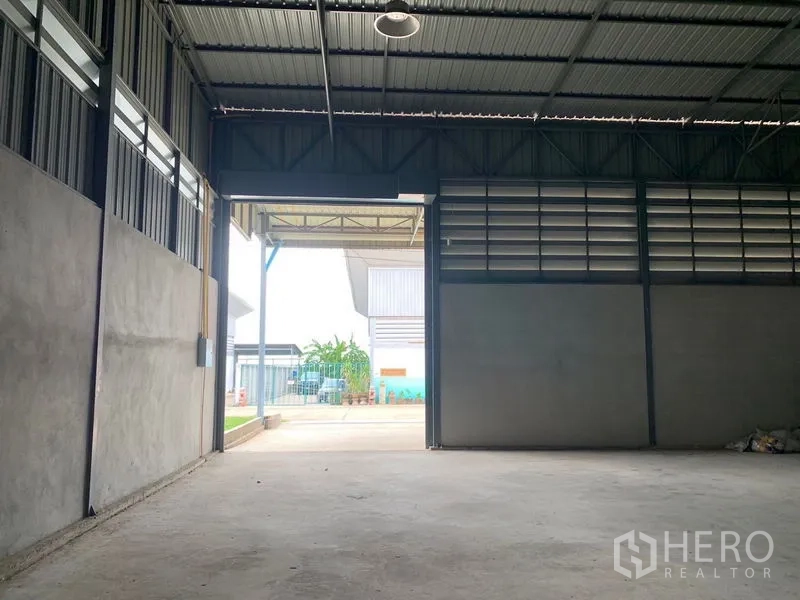 Warehouse for rent or sale in Sai Noi, Nonthaburi - Interior view toward the yard through a large shutter door and ventilated wall panels.