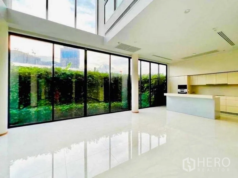 Detached House for sale in Huai Khwang, Bangkok - Bright double-height living area with floor-to-ceiling glass and a modern island kitchen facing a hedged garden.