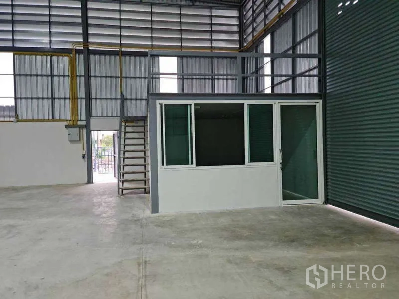 Warehouse for rent in Bang Bua Thong, Nonthaburi - Interior with glass-front office room and mezzanine platform next to a tall roller door.