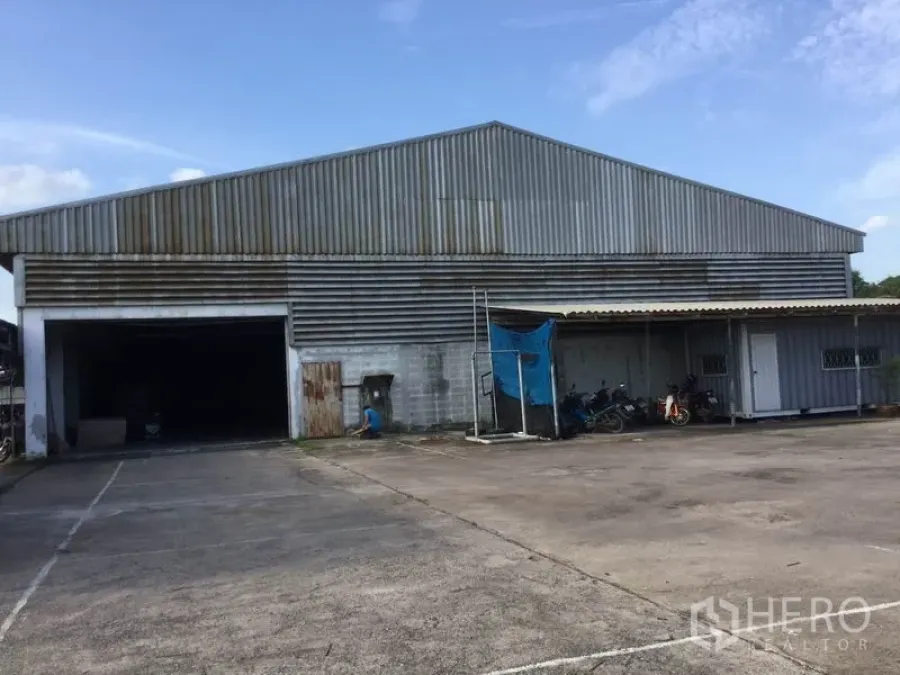 Warehouse for sale in Sai Noi, Nonthaburi - Front view of a large steel‑clad warehouse with a wide loading entrance and a small attached office under a canopy.