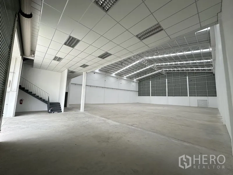 Factory for rent in Pathum Thani - Open warehouse interior with steel roof trusses, loading shutter, and mezzanine stair.