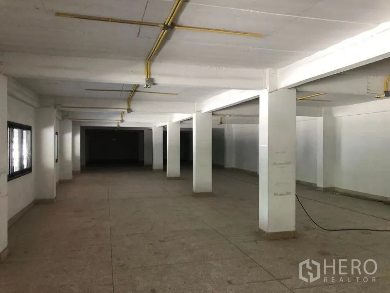 Warehouse for rent in Nonthaburi - Ground-floor office or storage area with low ceiling, white columns, windows, and exposed utility conduits.