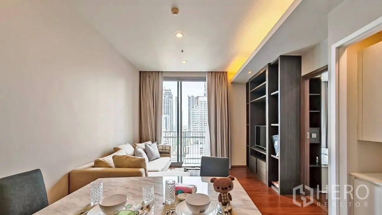Condominium for rent in Watthana, Bangkok - Open-plan living and dining room with an L-shaped sofa, built-in TV wall, wood flooring, and sliding glass door to a balcony with Bangkok skyline views.