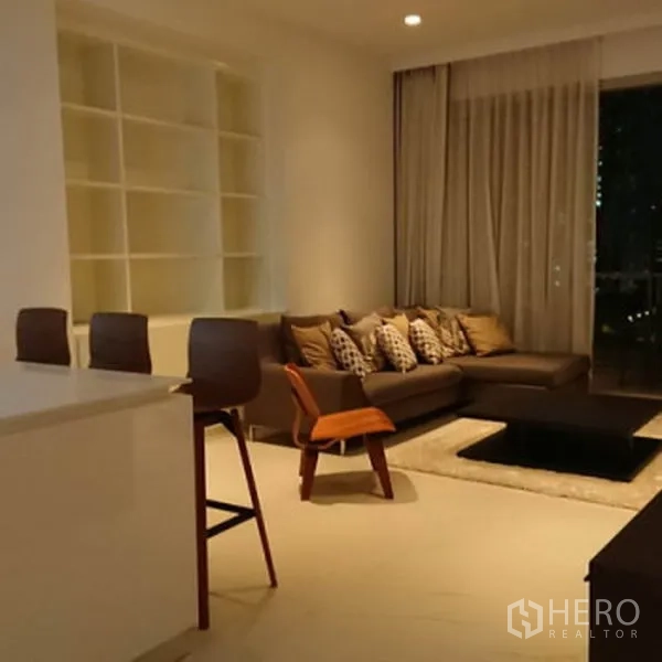 Condominium for rent in Pathum Wan, Bangkok - Warmly lit living room with L-shaped sofa, built-in shelving and floor-to-ceiling curtains opening to a balcony.