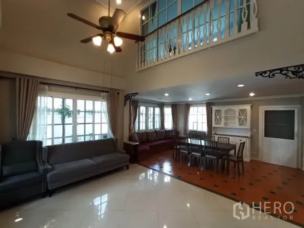 Detached House for rent in Mueang Samut Prakan, Samut Prakan - Double-height living room with ceiling fan, large windows, sofas and adjoining dining area with classic railing.