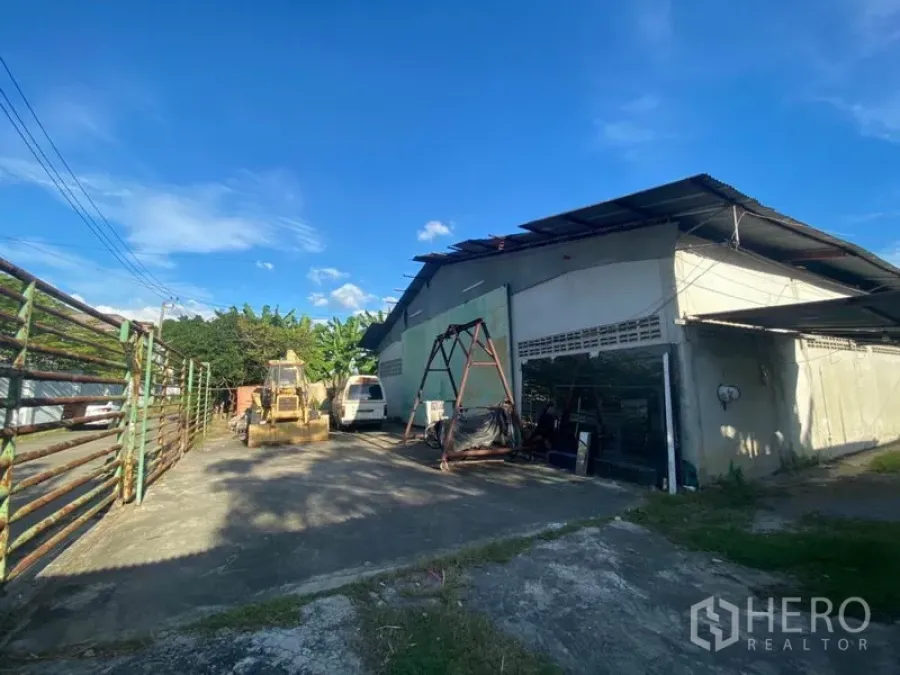 Warehouse for rent in Suan Luang, Bangkok - Warehouse exterior in Suan Luang with wide concrete yard, metal roof, machinery and van parking under a blue sky.