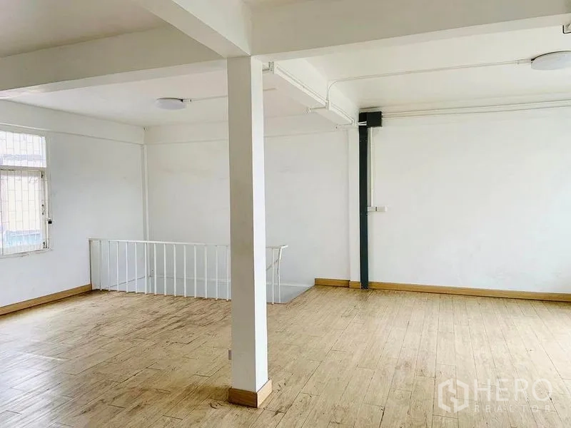 Shophouse for rent or sale in Khlong Toei, Bangkok - Spacious upper-floor room with wood-look flooring, white walls, a central column and a window beside the stairwell railing.