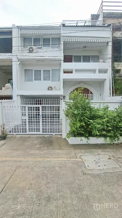 Townhouse for rent in Watthana, Bangkok - Front façade of the townhouse with white fence, balconies and lush shrub beside the gate.