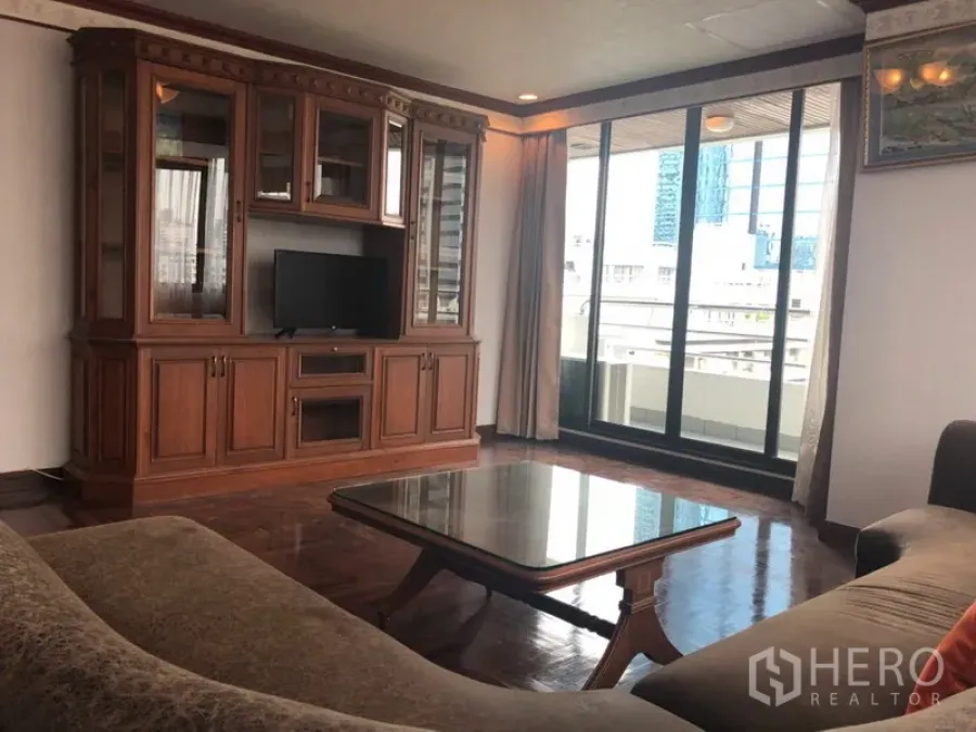Condominium for rent in Khlong Toei, Bangkok - Bright lounge with built-in wooden TV cabinet and sliding glass doors opening to a city-view balcony.