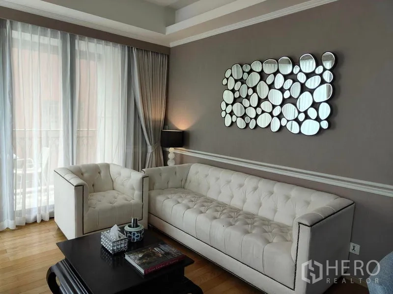 Condominium for rent or sale in Pathum Wan, Bangkok - Bright living room with tufted white sofa set, coffee table, and floor-to-ceiling curtains on hardwood floors.