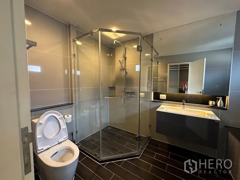 Detached House for rent or sale in Lam Luk Ka, Pathum Thani - Modern bathroom with glass shower enclosure, vanity and toilet on dark floor tiles.