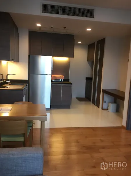 Condominium for rent in Khlong Toei, Bangkok - Modern kitchen with dark cabinetry, under-cabinet lighting, stainless refrigerator and a wooden dining table.