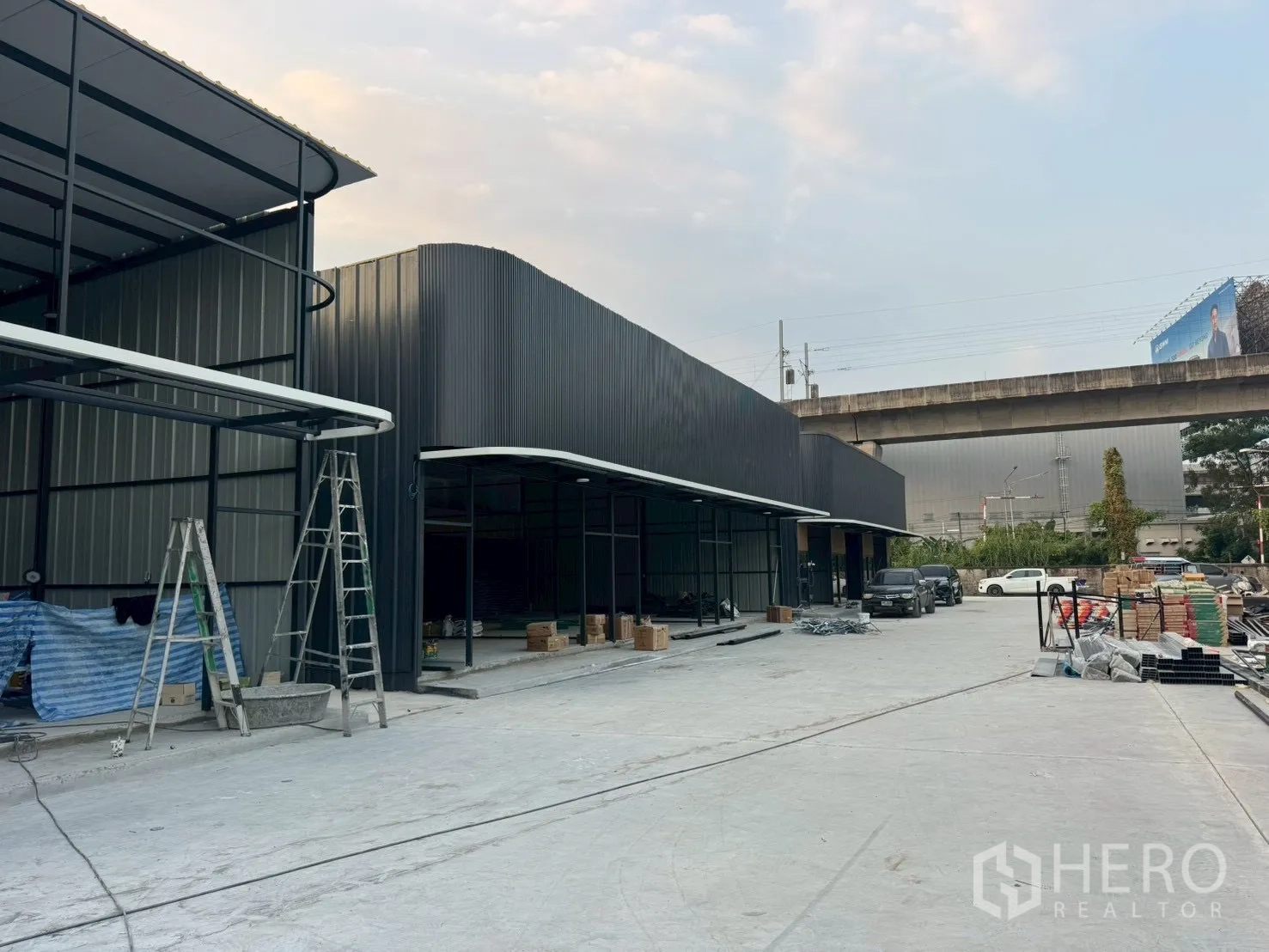 Warehouse for rent in Suan Luang, Bangkok - Row of steel warehouses under construction with ladders and a spacious yard beside an elevated rail line.