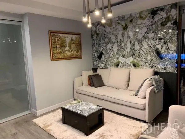 Condominium for sale in Khlong Toei, Bangkok - Modern living room of a Khlong Toei condo with beige sofa, statement marble accent wall, pendant lights and a glass sliding door.