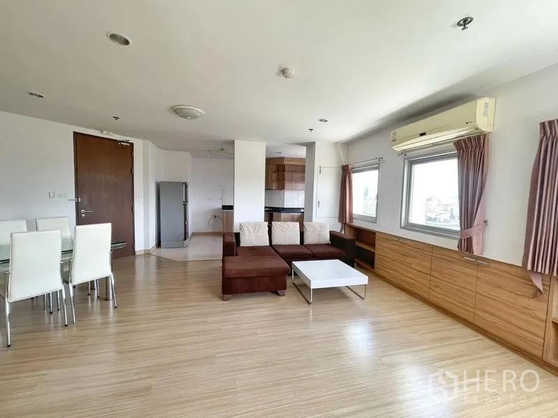 Condominium for rent in Khlong Toei, Bangkok - Open-plan living and dining area with L-shaped sofa, wood floors, built-in cabinets and large windows in a Khlong Toei condo.