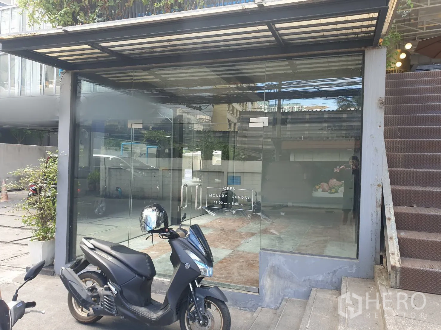 Retail Space for rent in Khlong Toei, Bangkok - Compact storefront with clear glass doors under a metal canopy next to stairs, with scooters parked in front.