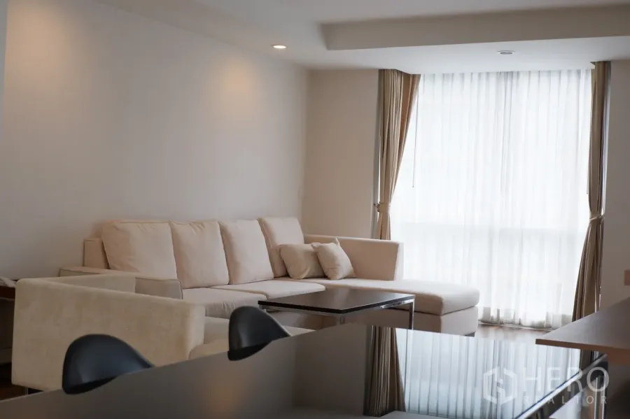 Condominium for rent in Pathum Wan, Bangkok - Cozy living room with large beige sectional, coffee table, and corner windows with sheer curtains.