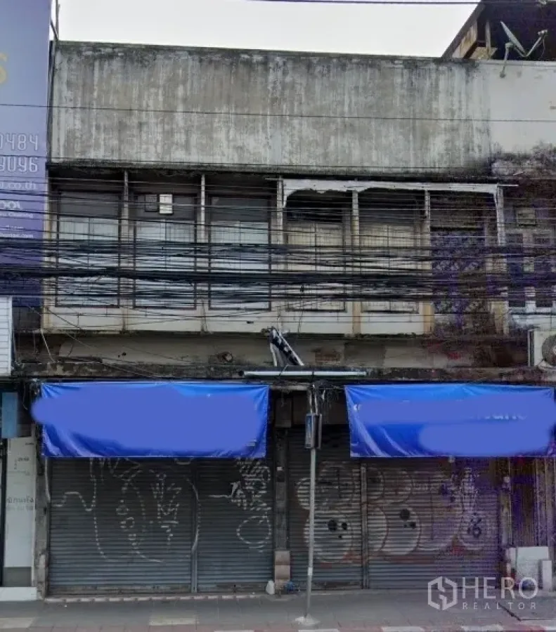 Shophouse for sale in Watthana, Bangkok - Street-facing shophouse facade with blue awnings, metal roller shutters, and upper barred windows in Watthana, Bangkok.