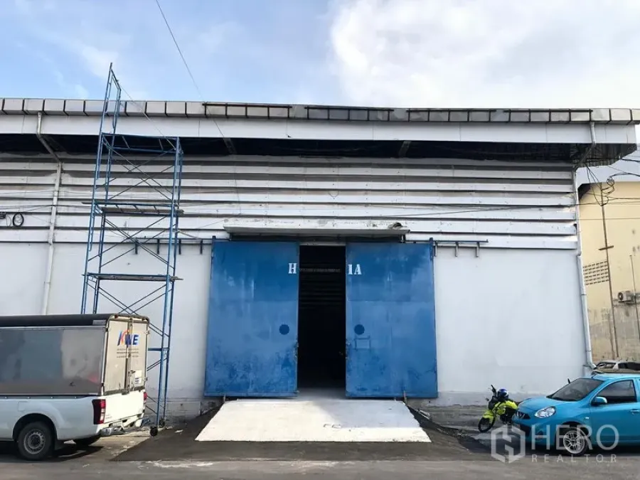 货栈 出租在Bang Phli, Samut Prakan - 仓库外立面，白色墙体与蓝色推拉大门，带混凝土装卸坡道，前方可停车