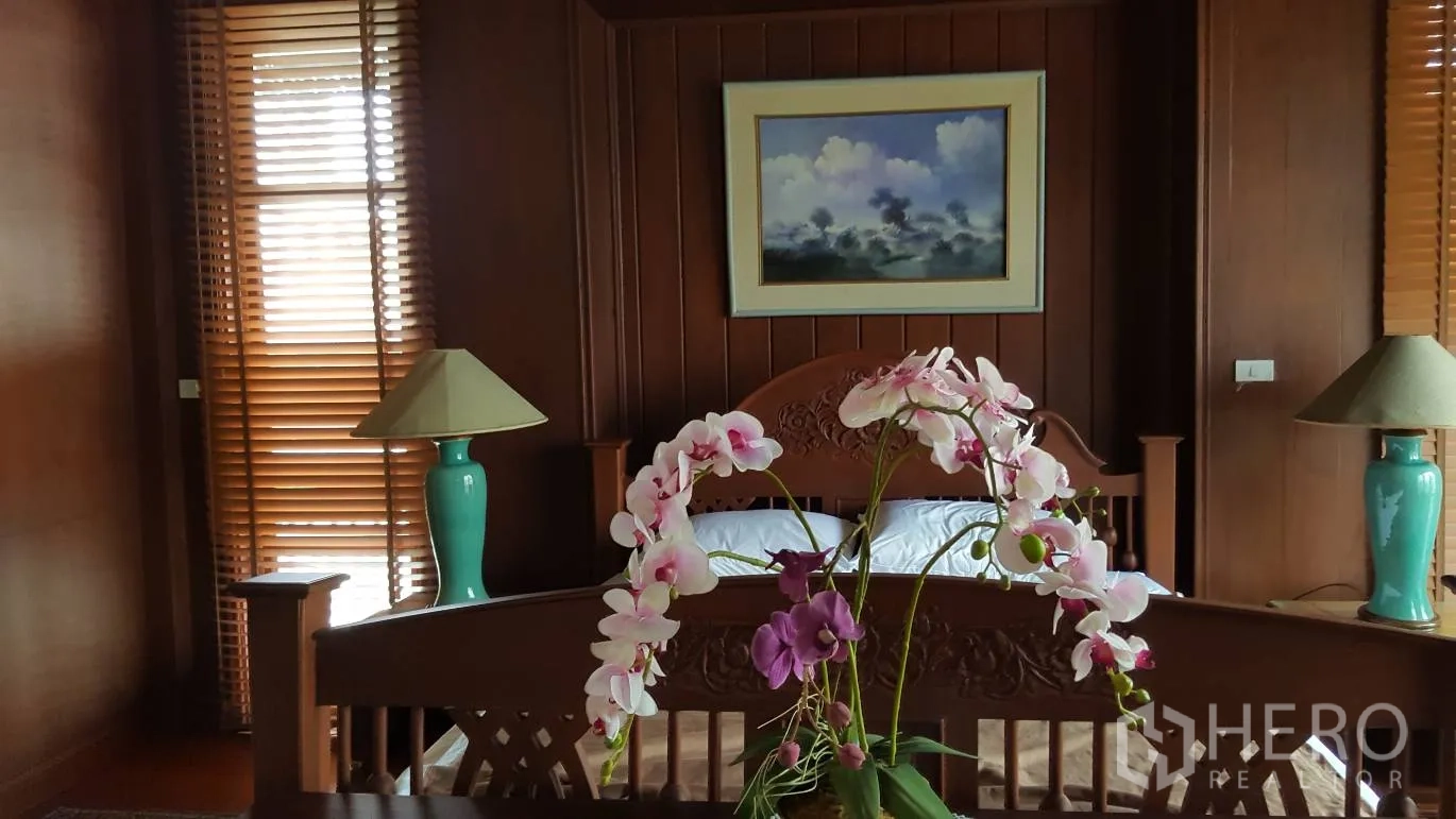 Detached House for rent in Khlong Toei, Bangkok - Warm teak bedroom with a carved wooden bed, bedside lamps, window blinds and an orchid centerpiece.