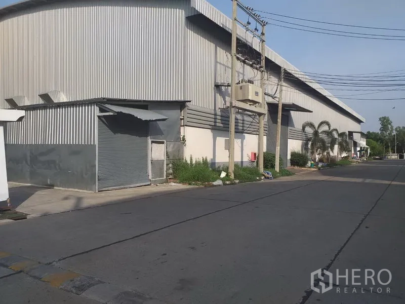 Factory for rent in Phra Nakhon Si Ayutthaya - Side view of a corrugated metal warehouse with a roller shutter door and utility lines along the street.
