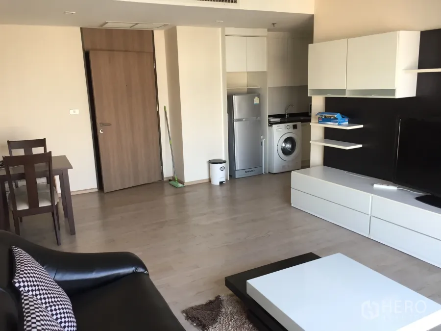 Condominium for rent or sale in Khlong Toei, Bangkok - Open-plan living room with dining set, TV console, and compact kitchenette with fridge and washing machine.