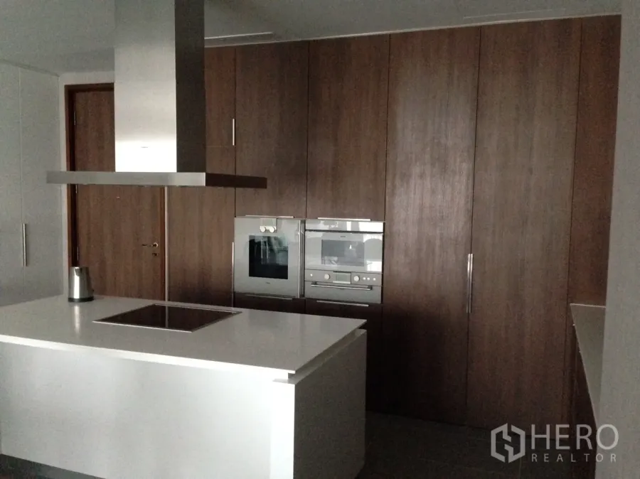 Condominium for rent or sale in Pathum Wan, Bangkok - Modern kitchen with a white island, induction hob, stainless-steel hood and built-in ovens set against warm wood cabinetry.