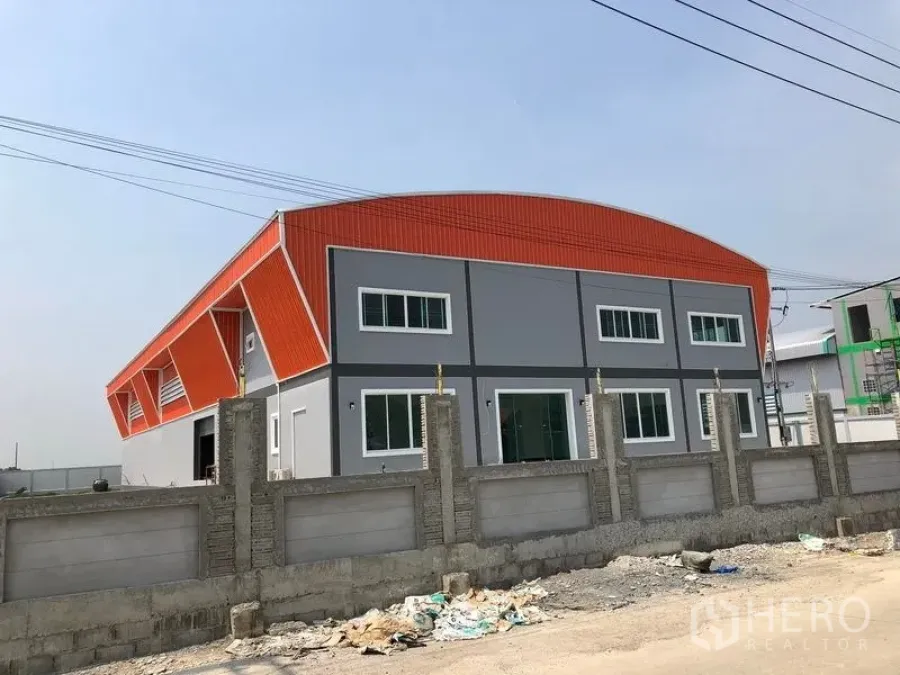 Factory for sale in Mueang Samut Sakhon, Samut Sakhon - Modern two-storey factory with orange curved roof and grey facade behind a concrete perimeter wall in Khok Kham.