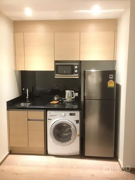 Condominium for rent in Khlong Toei, Bangkok - Compact modern kitchenette with light wood cabinets, black stone countertop, microwave, washing machine, and stainless refrigerator.