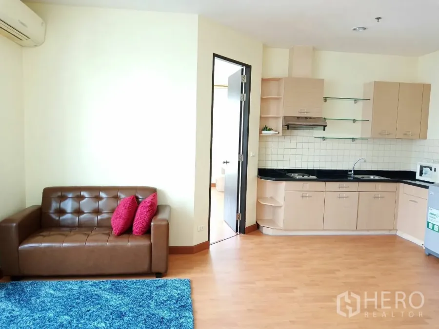 Condominium for rent in Khlong Toei, Bangkok - Open-plan living area with a brown leather sofa, blue rug, and light-wood kitchen with black countertops and tile backsplash.