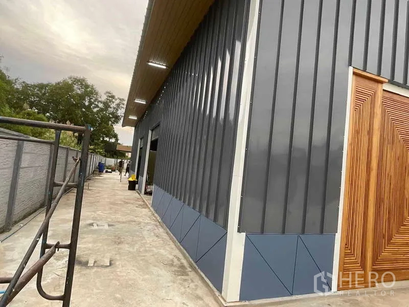 Warehouse for rent or sale in Nong Chok, Bangkok - Side view of a steel‑clad warehouse with canopy lighting and wooden doors along a concrete driveway.