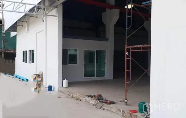 Factory for rent in Krathum Baen, Samut Sakhon - Exterior of the factory with white façade, glass-fronted office, covered loading bay and construction tools on the apron.