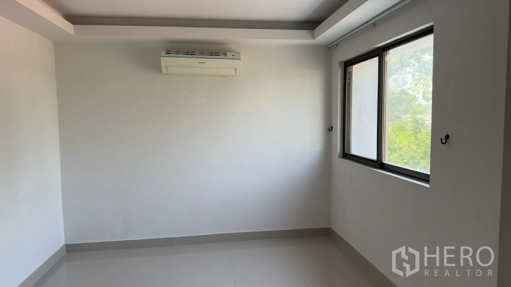 Townhouse for rent in Watthana, Bangkok - Cozy bedroom with a wall air conditioner and two windows overlooking greenery.