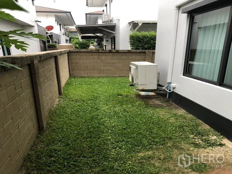 Detached House for rent in Bang Kapi, Bangkok - Side yard with grass lawn, brick privacy wall and exterior A/C unit.