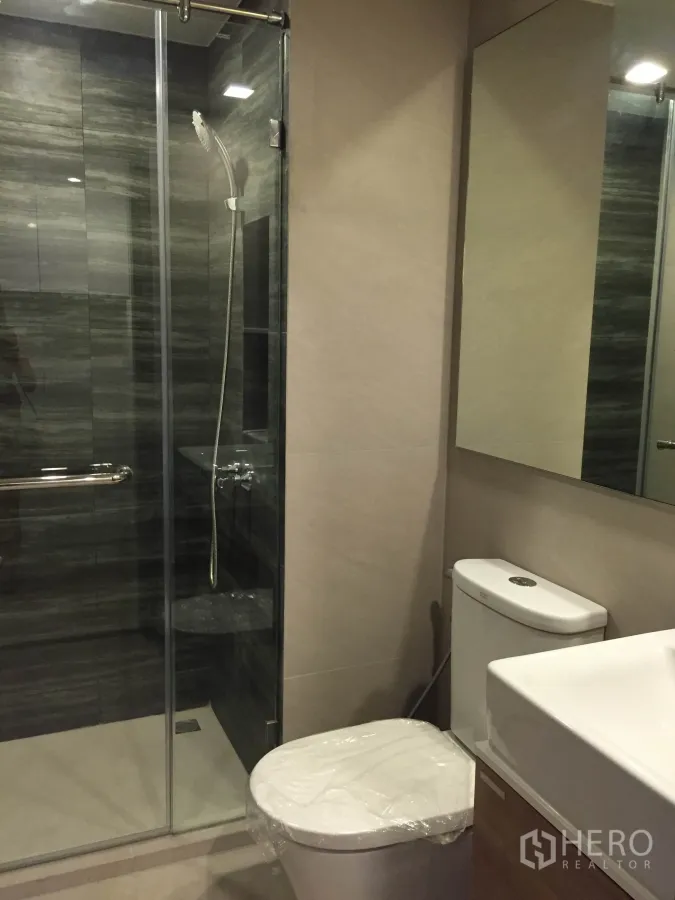 Condominium for rent in Pathum Wan, Bangkok - Modern bathroom with glass-enclosed shower, handheld showerhead, large mirror and toilet in neutral tones.
