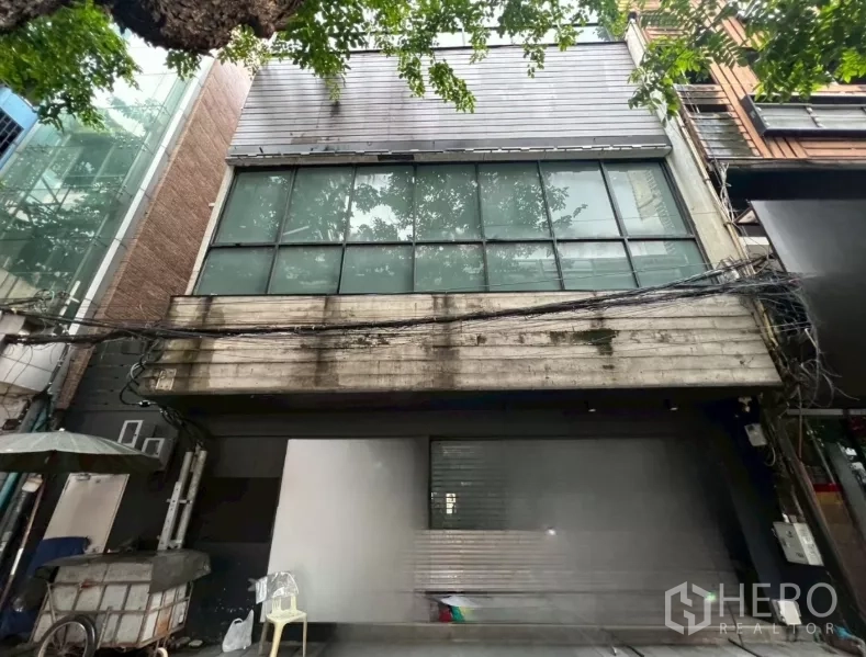 店屋 出租在Bang Rak, Bangkok - 临街商铺外立面，宽大玻璃窗与金属饰面，树荫掩映