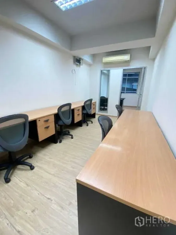 Office Space for rent in Khlong Toei, Bangkok - Long workspace with multiple built-in desks, task chairs, wall-mounted air conditioner and bright fluorescent lighting.