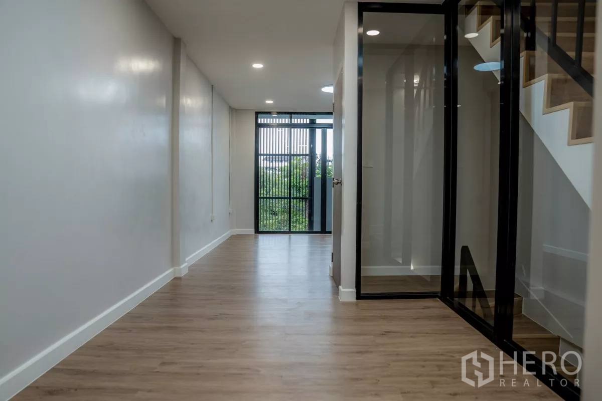 Townhouse for sale in Watthana, Bangkok - Long corridor with recessed lighting leading to a balcony and glass stair enclosure.