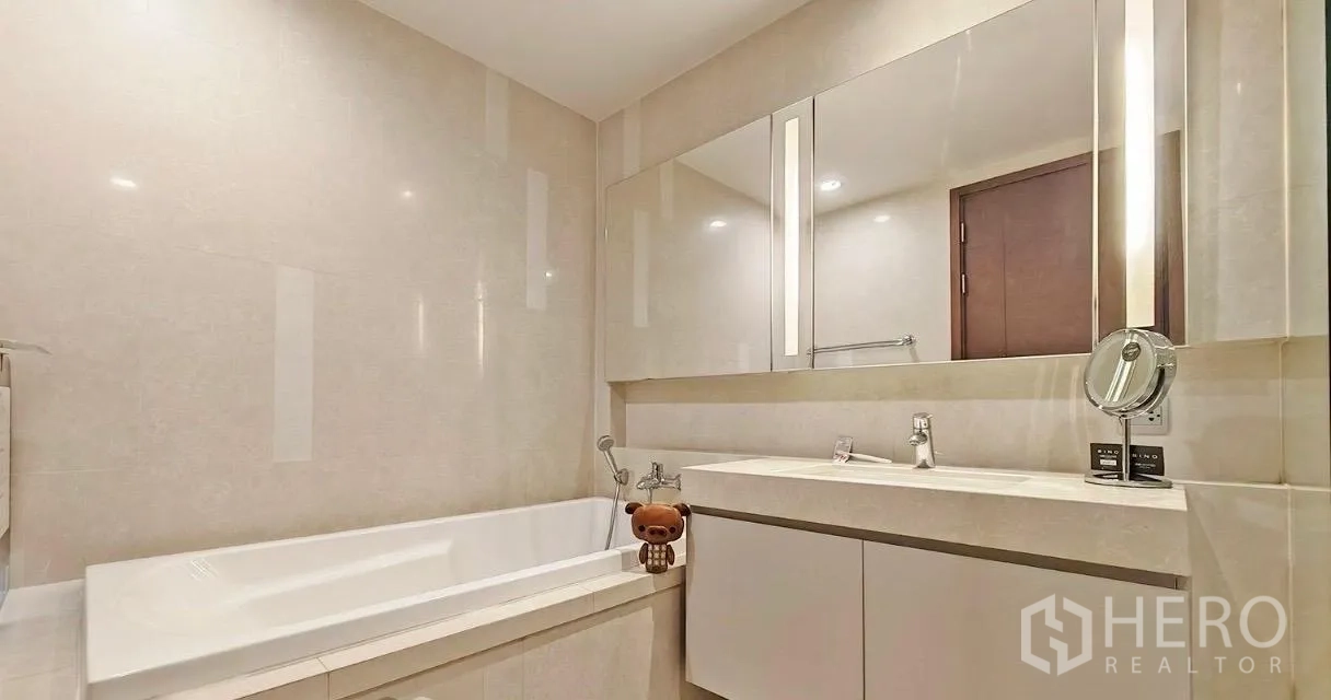 Condominium for rent in Watthana, Bangkok - Contemporary bathroom with a long bathtub, wide mirror, vanity, and neutral stone tiles.