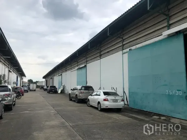 Warehouse for rent in Bang Phli, Samut Prakan - Row of warehouses with teal sliding doors and a wide driveway with parked vehicles for easy truck access.