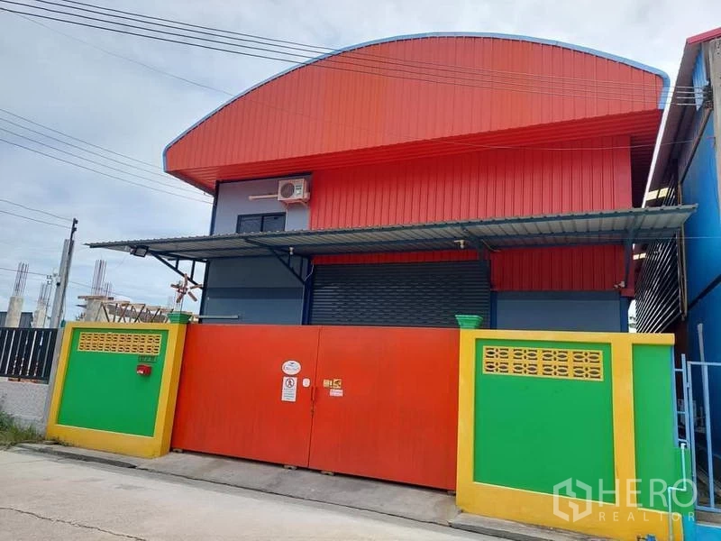 Warehouse for rent or sale in Bang Bua Thong, Nonthaburi - Colorful steel-clad warehouse with arched red roof, wide roller shutter, and secure front gate.