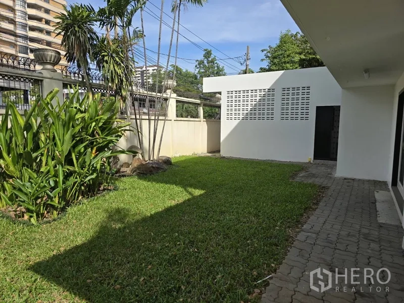 Detached House for rent in Watthana, Bangkok - Side lawn with walkway, palms and a white utility building inside the walled compound.