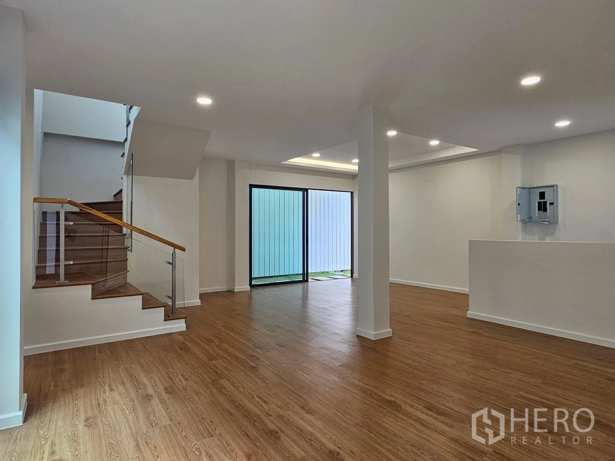 Detached House for sale in Lat Phrao, Bangkok - Open-plan living area with wood-style flooring, recessed lights, and a glass-railed staircase beside sliding doors to a courtyard.