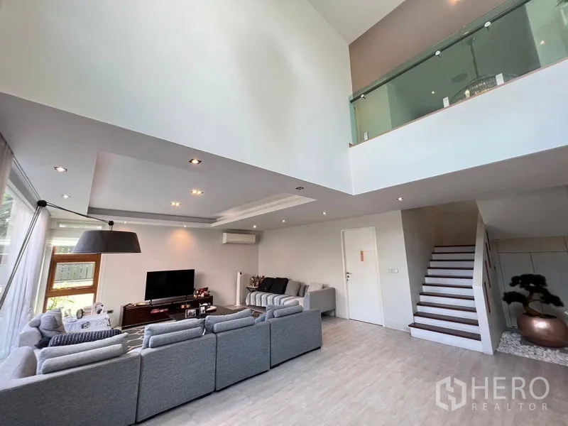 Detached House for sale in Chatuchak, Bangkok - Double-height living room with L-shaped sofa, TV console, recessed lighting and mezzanine with glass balustrade.