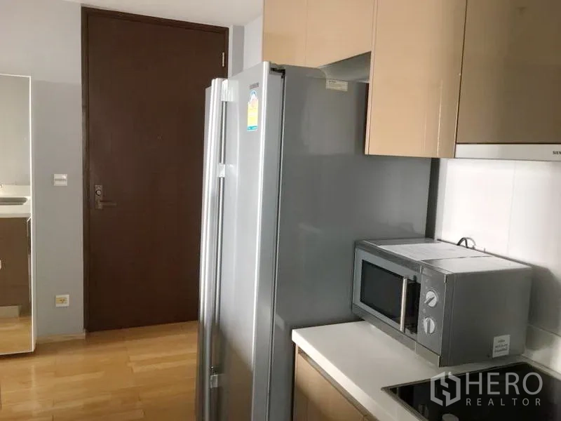 Condominium for rent in Khlong Toei, Bangkok - Compact modern kitchenette with stainless refrigerator, microwave, electric cooktop and glossy upper cabinets.