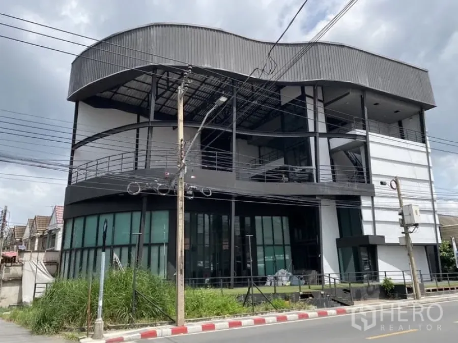 Showroom for rent in Lat Phrao, Bangkok - Four-storey showroom building in Lat Phrao with curved metal roof and full-height glass frontage on a street corner.